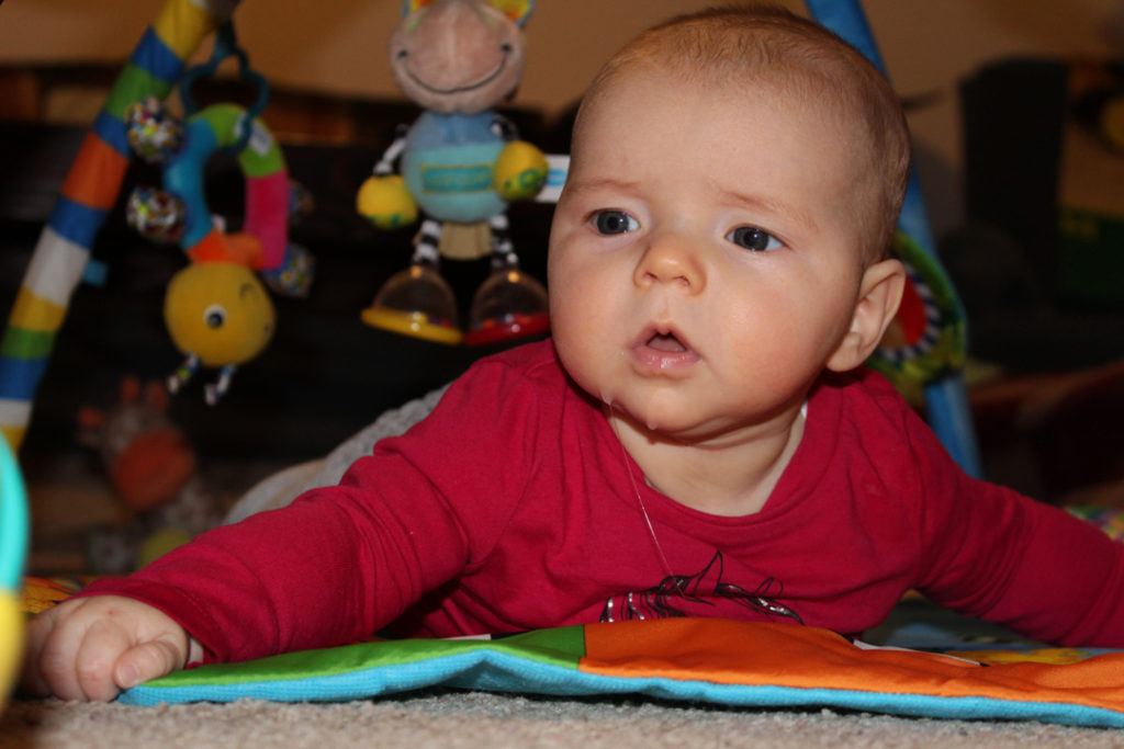 3 month old doing tummy time. Her hands are spread, her head is lifted, and her head and eyes are looking to the right at an object barely shown in the corner of the image. 