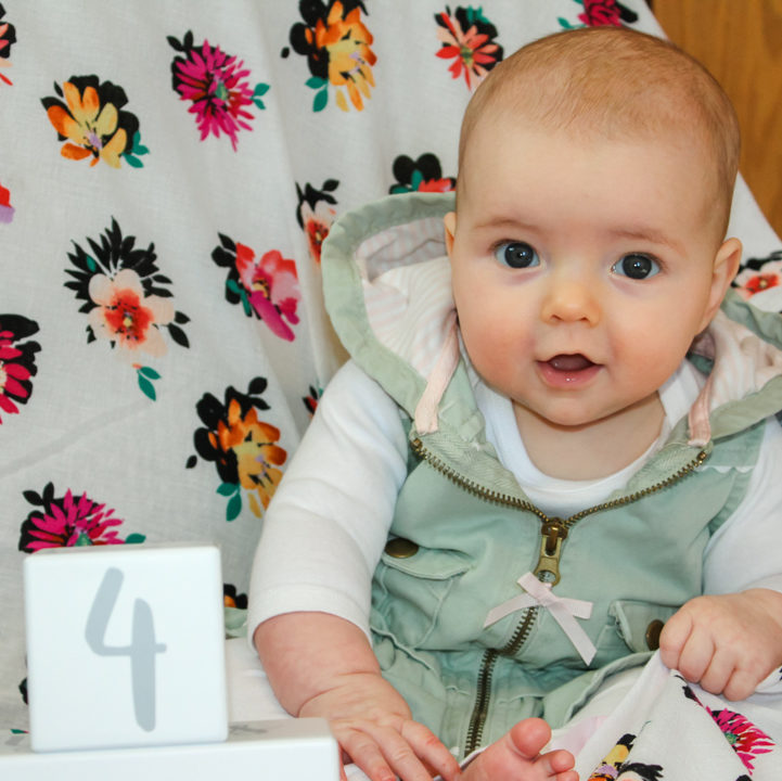 4 month old baby girl in a white long sleeve onesie with a light green vest on. She is sitting in the corner of a rocking chair covered in a flower blanker. There is a white block with a gray number four on it in front of her. The baby is slightly leaning forward grasping the blanket with her left hand looking at the camera with both eyes moving in alignment. 