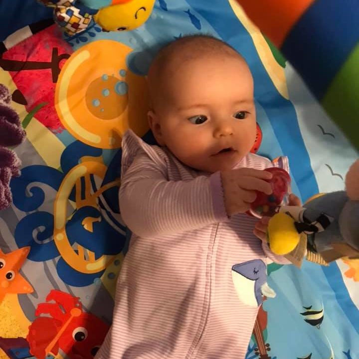 2 month old baby laying on her back in on a colorful play gym. her eyes are working together to inspect the bottom of a hanging toy that is above her. She is holding one foot of the toy with both hands. The baby's eyes do not have perfect eye movement alignment yet.