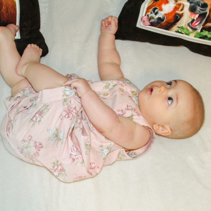 6 month old baby girl laying on a white blanket. To her right are two farm animal pillows. She is twisted slightly to her right with her eyes cut to the side and up to look at one of the pillows. Her head is not fully turned. The baby's eye movements are developing on track.
