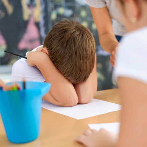 little boy at school with his head down and his arms and hands covering his face and ears ontop of his paper