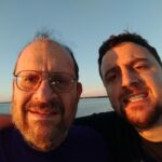 Arthur Henick and his adult son Ian smile at the camera. In the background is a body of water and a dark blue sky.